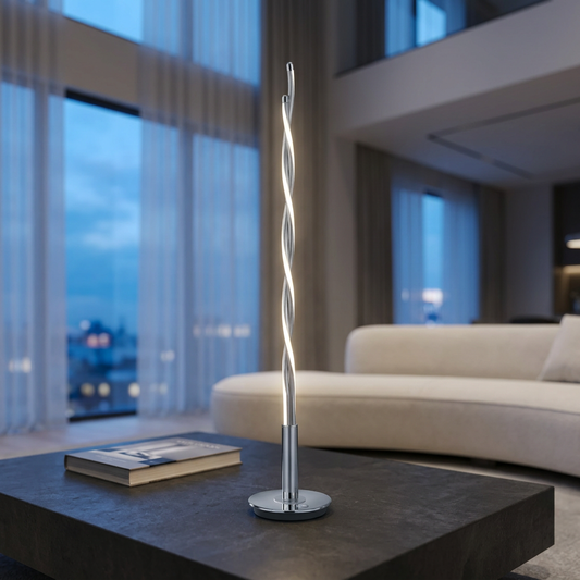 Eingeschaltete schlanke LED-Tischleuchte TWIST mit gedrehten Lichtbändern und Chromfuß auf einem dunklen Steintisch vor einem hellen Sofa