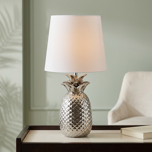 Tischleuchte ANANAS mit silberfarbenem Keramikfuß in Ananasform und weißem Textilschirm auf einer eleganten Konsole vor salbeigrüner Wand und cremefarbenem Sessel.