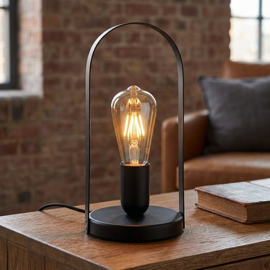 Schwarze FARO-Metall-Tischleuchte mit offenem Rundbogen und leuchtendem Filament-Leuchtmittel auf einer Eichenkonsole vor einer Backsteinwand.