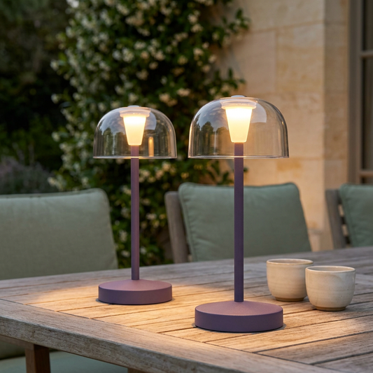 Zwei brombeerfarbene GOMBA-Akku-Tischleuchten mit transparenten Kuppelschirmen und warmem Licht auf einem Teakholztisch auf der Terrasse.