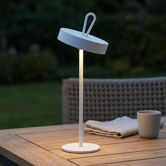 Eingeschaltete weiße LED-Akku-Tischleuchte ELLA mit geneigtem rundem Leuchtenkopf auf einem Teakholztisch auf einer Terrasse am Abend